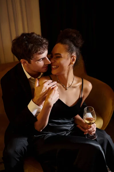 handsome man in suit embracing happy african american woman with glass of wine and sitting on sofa