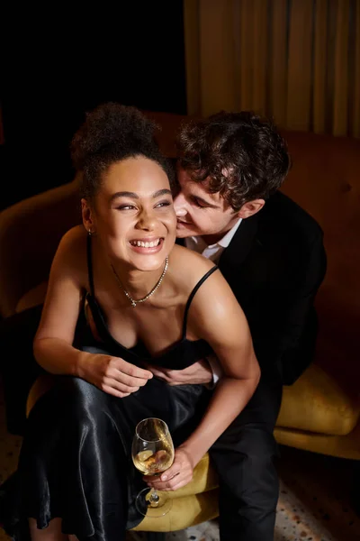 handsome man in suit embracing joyful african american woman with glass of wine and sitting on sofa