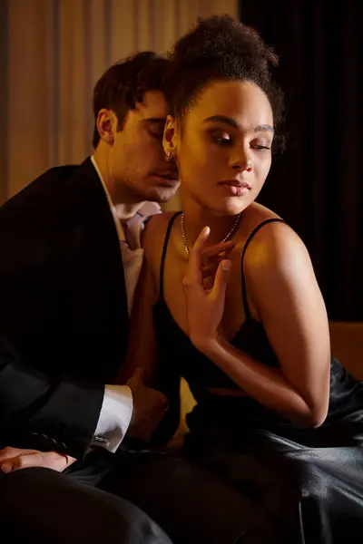 good looking man in suit seducing pretty african american woman in black dress and sitting on sofa