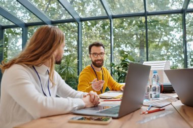 two cheerful businessmen in casual vivid attires discussing their important startup at table clipart