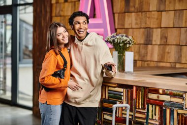 happy diverse couple, dressed in casual clothes standing happily surrounded by shelves of books clipart