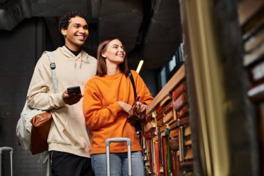 Akıllı telefonlu mutlu ve çeşitli çiftler yurtta kitaplığın yanında neşeli bir anı paylaşıyorlar.