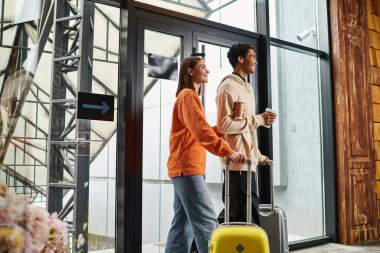 Yolculuk valizli mutlu çift gülümseyerek modern bir otele giriyor, ellerinde kahve var.