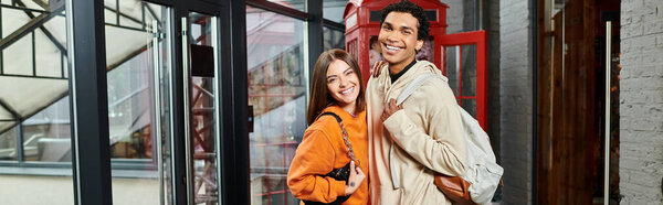 Cheerful diverse couple stands by a red phone booth in modern hostel, ready for travel banner