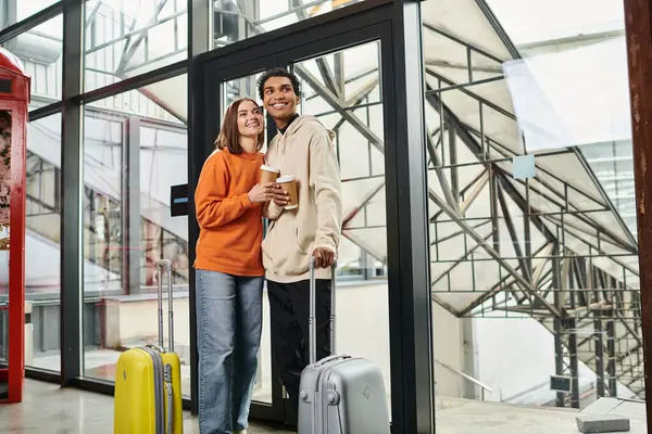 Young diverse couple with travel luggage smiling and entering a modern hostel, holding coffee to go