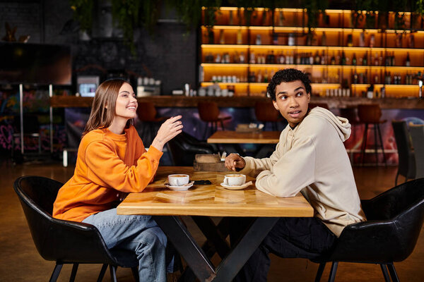 cheerful woman showing something to black man while sitting together in a cozy cafe, cups of coffee