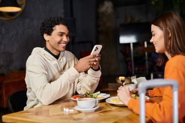 Bir kadın karşısında oturmuş kahvaltı ederken mutlu siyah adam akıllı telefon kullanıyor.