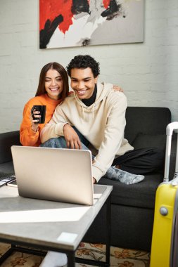 Mutlu ve çeşitli çiftler kanepede oturup dizüstü bilgisayar ve kahve kullanırken samimi bir anı paylaşıyorlar.