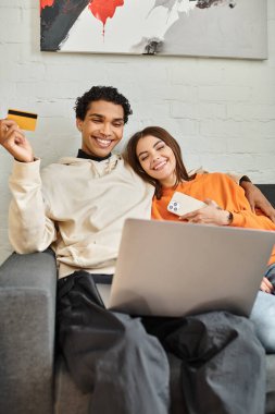 İnternetten alışveriş yapan mutlu çiftler, ellerinde kredi kartı ve pansiyondaki kanepede dizüstü bilgisayarları var.