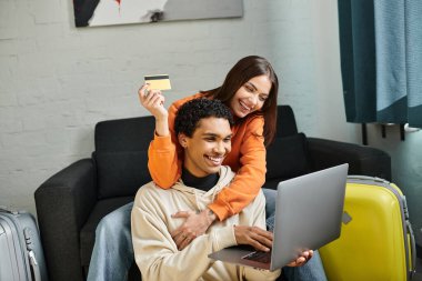 İnternetten alışveriş yapan mutlu çift, ellerinde bir kredi kartı, bavullarının yanında dizüstü bilgisayarları var.