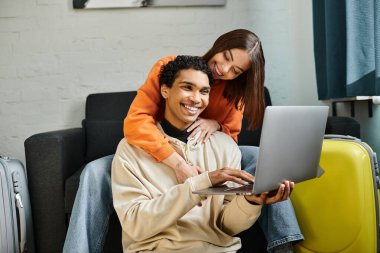 Çevrimiçi alışveriş yapan neşeli çift, ellerinde bir kredi kartı, bavullarının yanında dizüstü bilgisayarları var.