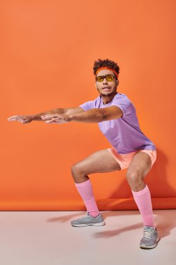 Güneş gözlüklü ve spor kıyafetli genç Afro-Amerikan sporcusu turuncu arka planda çömelmiş.