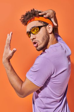 Gözlüklü ve bantlı Afro-Amerikan adam turuncu arka plandaki kameraya bakıyor.
