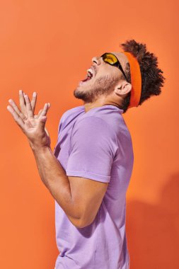 expressive african american man in eyeglasses and headband screaming on orange background clipart