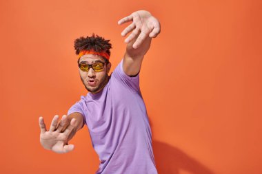 Gözlüklü ve saç bantlı komik ve anlamlı Afro-Amerikalı adam turuncu arka planda el kol hareketi yapıyor.