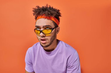 annoyed african american man in eyeglasses and headband rolling eyes on orange background clipart