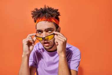 annoyed african american man in eyeglasses and headband rolling eyes on orange background, emotion clipart