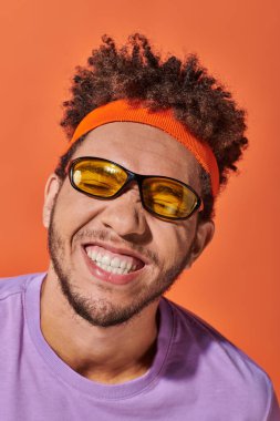 happy african american man in eyeglasses and headband grinning on orange background, grimace clipart