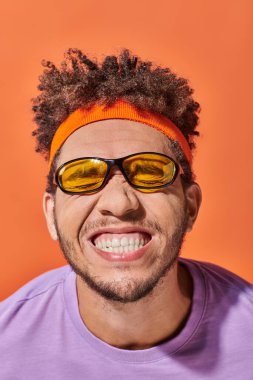 funny african american fella in eyeglasses and headband grinning on orange background, grimace clipart