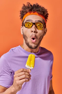 Şok olmuş, güneş gözlüklü ve saç bantlı Afro-Amerikan adam portakal arkaplanda meyveli dondurma tutuyor.