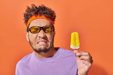 Güneş gözlüğü ve saç bandı takmış, portakallı dondurmaya bakan hoşnutsuz Afro-Amerikan adam.