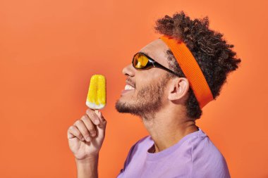 Güneş gözlüklü ve bantlı mutlu Afro-Amerikan adam elinde dondurmayla turuncu arka planda.