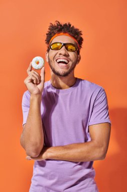 Güneş gözlüklü, turuncu arka planda lezzetli çörekler tutan mutlu, Afro-Amerikan bir adam.