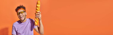 happy african american man in sunglasses holding fresh baguette on orange background, banner clipart