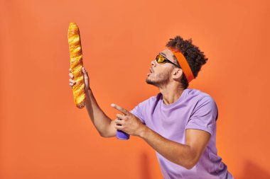 Güneş gözlüklü Afro-Amerikan adam turuncu arka planda taze ekmek ve sodaya bakıyor.