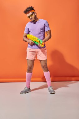young african american guy in sunglasses and summer outfit holding water gun on orange background clipart