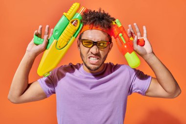 tensed african american man in sunglasses  holding water guns on orange background, playful clipart