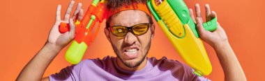 tensed african american man in sunglasses  holding water guns on orange background, banner clipart