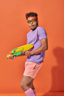 Kafası karışmış, güneş gözlüklü, turuncu arka planda su tabancası tutan Afro-Amerikan bir adam.