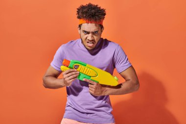 african american man in headband playing water fight with toy gun on orange background, grimace clipart