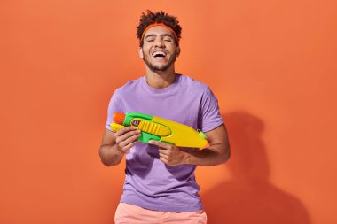 Kafasında saç bandı olan mutlu Afro-Amerikalı adam turuncu arka planda oyuncak tabancayla su savaşı yapıyor.