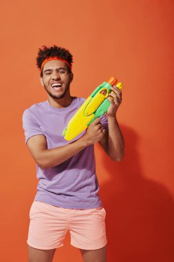 positive african american man in headband playing water fight with toy gun on orange background clipart