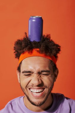 purple soda can on head of happy curly african american guy with headband on orange background clipart