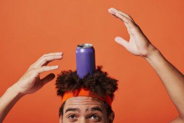 purple soda can on head of curly african american man with headband on orange background, gesture clipart