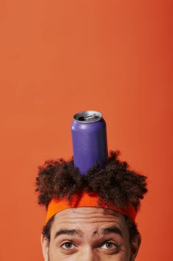 Turuncu arka planda saç bandı olan kıvırcık Afrikalı Amerikalı bir adamın kafasında mor soda kutusu var.