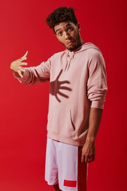 confused african american man in hoodie gesturing and looking at camera on red background, shadow clipart