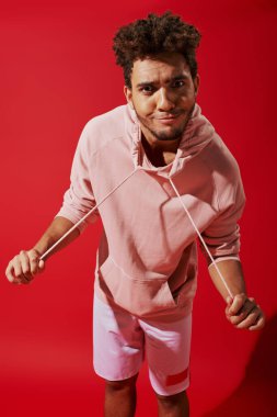 funny african american man pulling drawstrings of hoodie and looking at camera on red background clipart