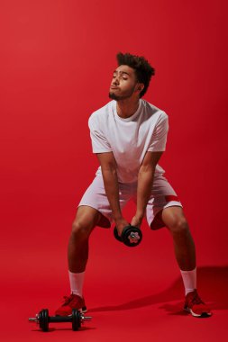 Kırmızı arka planda dambılla spor yapan komik Afro-Amerikan sporcusu.