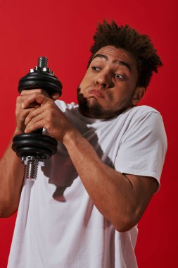 young african american man working out with heavy dumbbell and puffing cheeks on red background clipart
