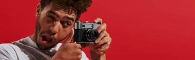 funny african american man looking at viewfinder while taking shot on red background, banner clipart