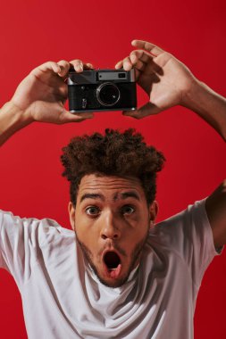amazed african american photographer with open mouth taking shot on retro camera on red background clipart