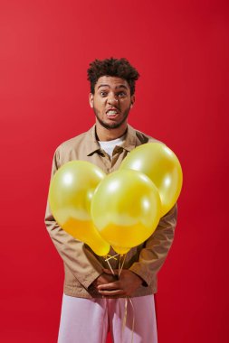 Beyaz ceketli komik Afro-Amerikan adam balon tutuyor ve kırmızı arka planda suratını buruşturuyor.