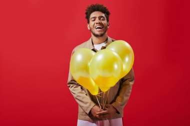 Bej ceketli neşeli Afro-Amerikan adam elinde balonlarla kırmızı arka planda gülüyor.