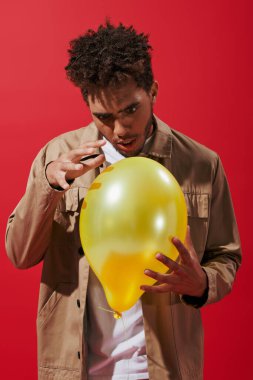 enchanted african american man in beige jacket looking at balloon on red background, amusement clipart