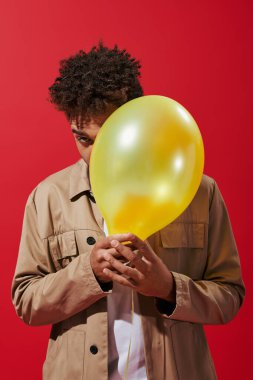 Kırmızı arka planda balonun arkasına saklanan bej ceketli Afro-Amerikan genç adam.