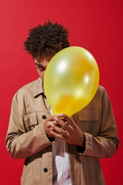 young african american man in beige jacket hiding behind balloon on red background, obscuring face clipart